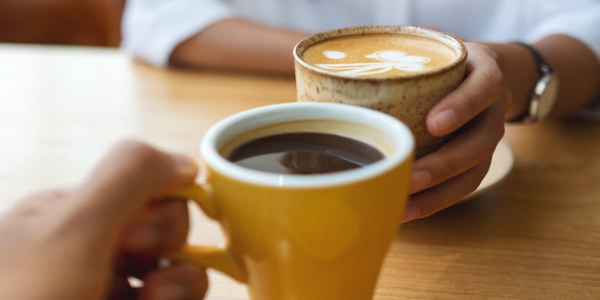 Two hands holding different coffee cups, one yellow and one with latte art, on a wooden table.