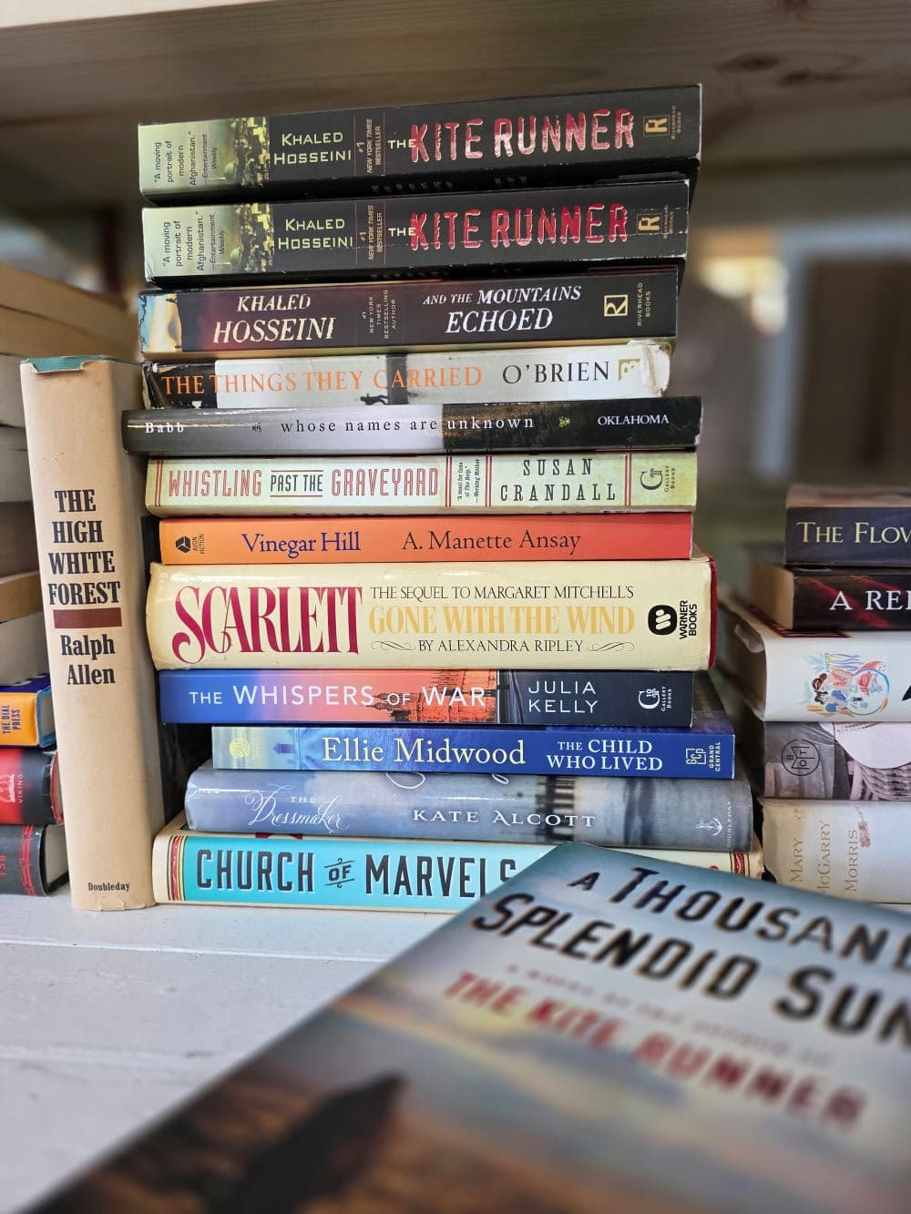 A stack of various books, including titles by Khaled Hosseini and others, displayed on a wooden shelf.