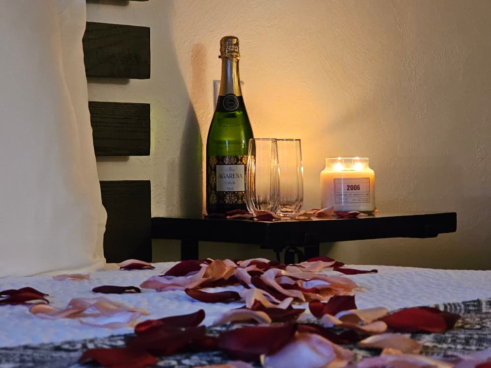 A bottle of champagne, two glasses, a lit candle, and scattered rose petals decorate a bedside table.