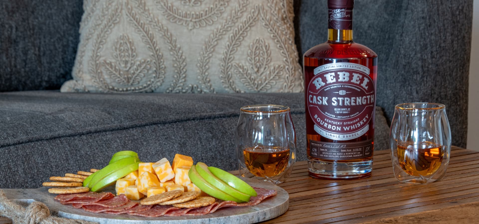 A bottle of Rebel Cask Strength bourbon sits beside two glasses and a platter of crackers, cheese, salami, and sliced apples on a wooden table.