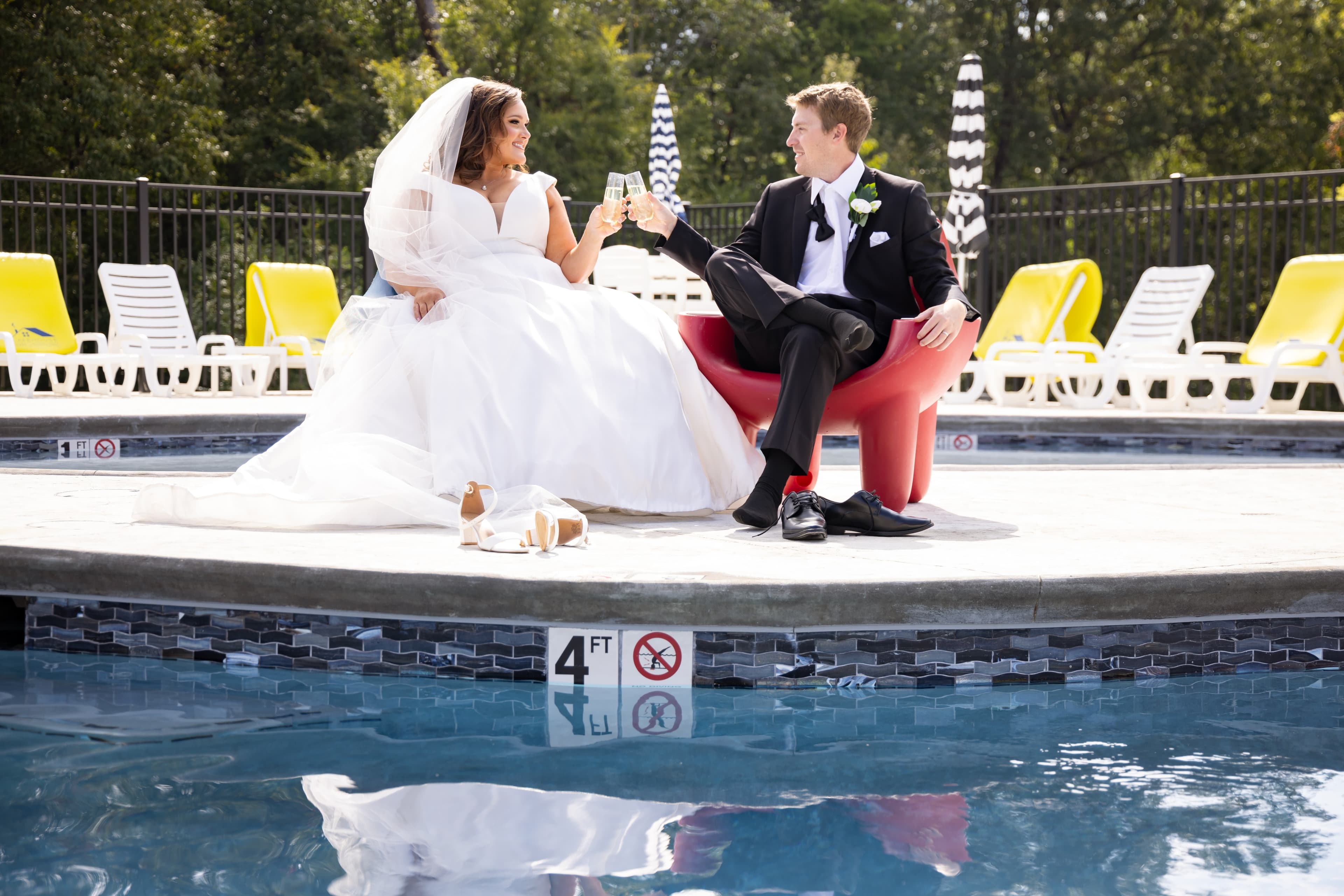A newlywed couple clinks glasses while seated by a poolside.