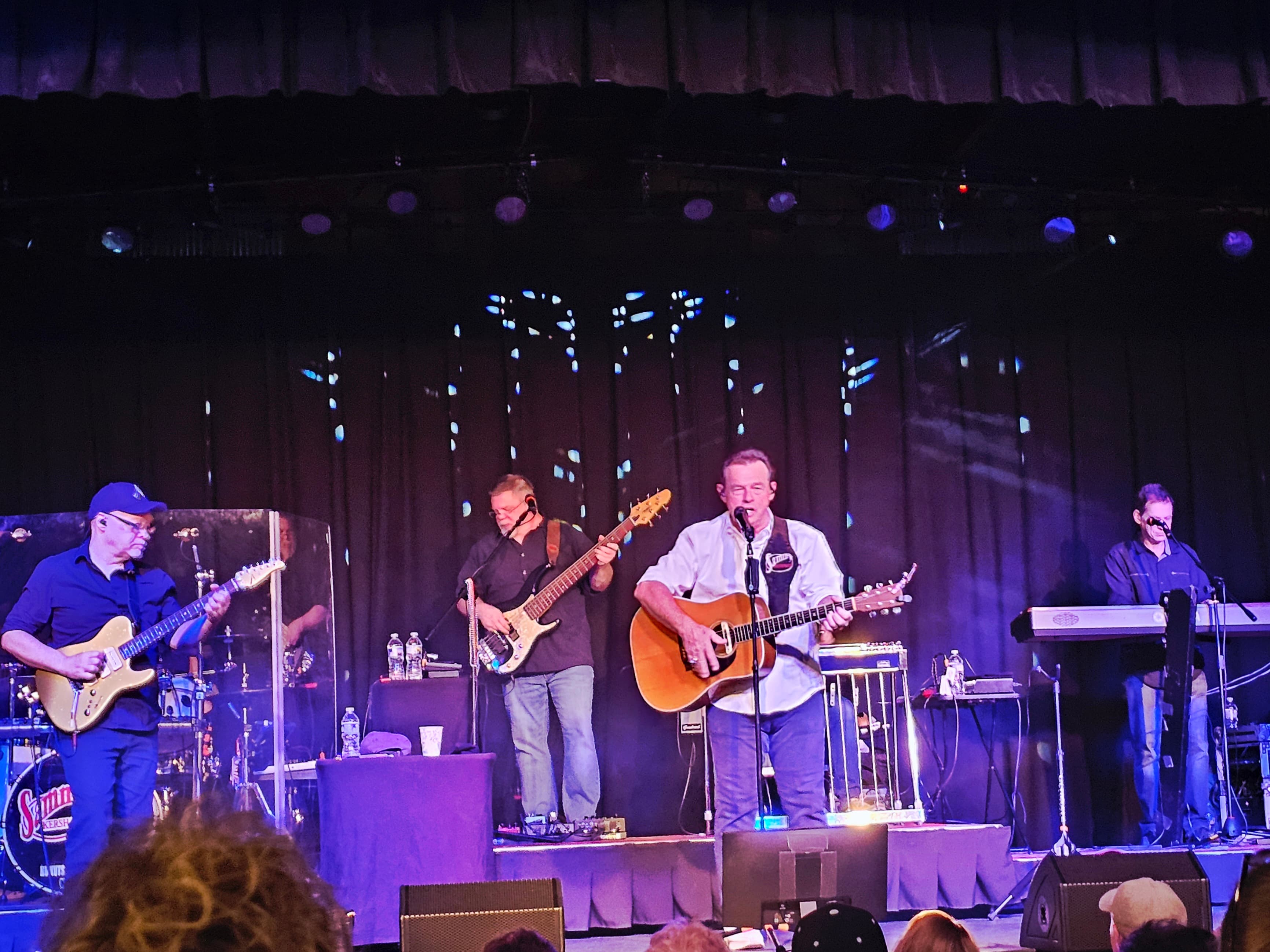 A live band performs on stage with an acoustic guitarist and backing musicians.