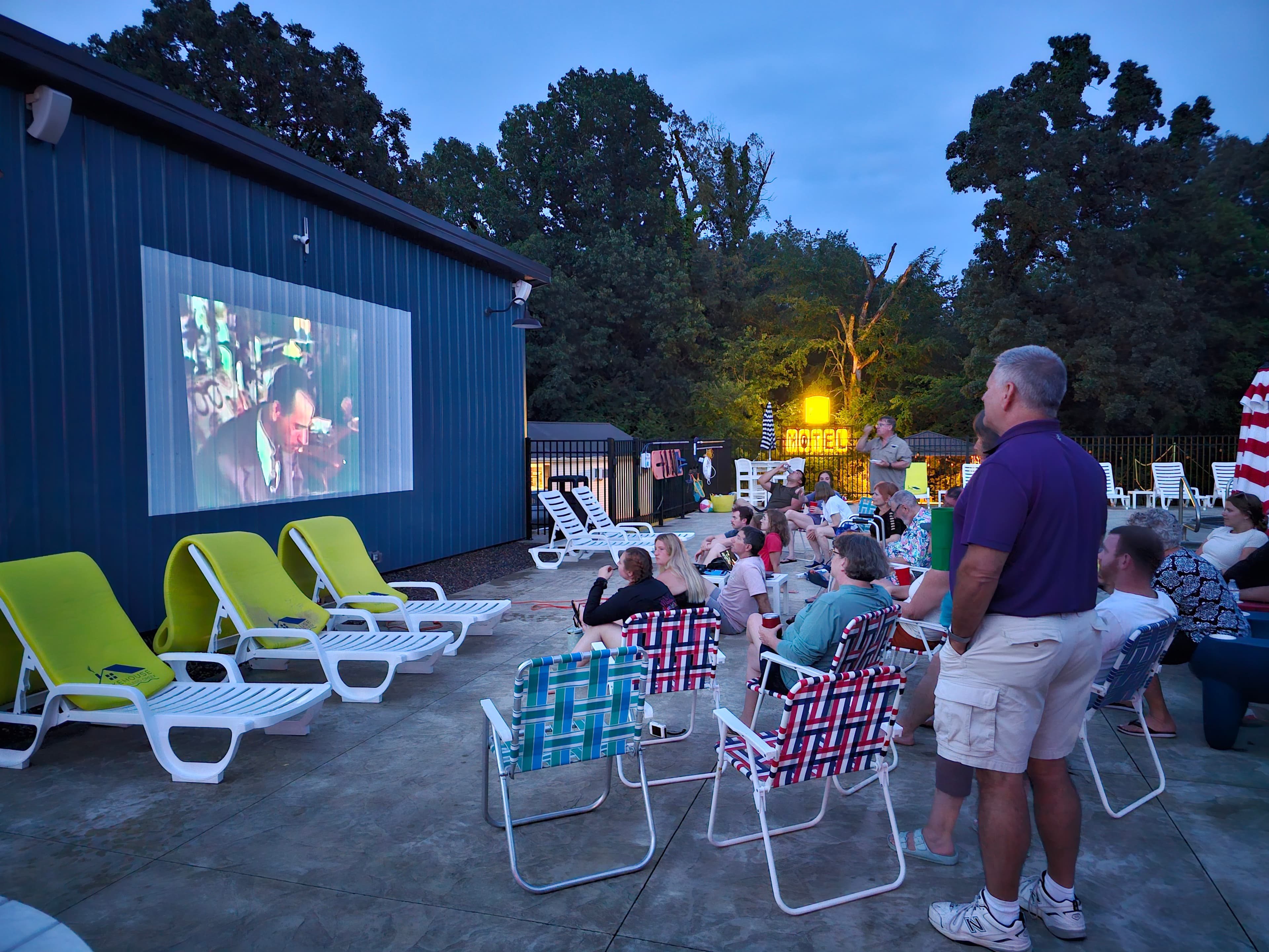 A group of people sits on lounge chairs watching a movie projected on an outdoor screen at dusk.