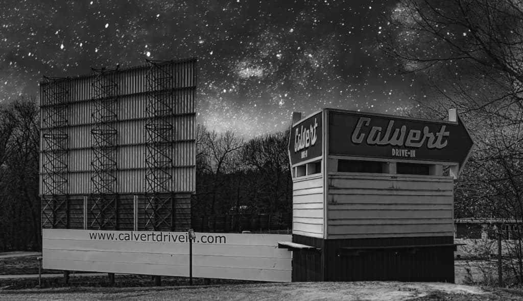 Black and white image of the Calvert Drive-In sign and a large empty movie screen under a starry sky.