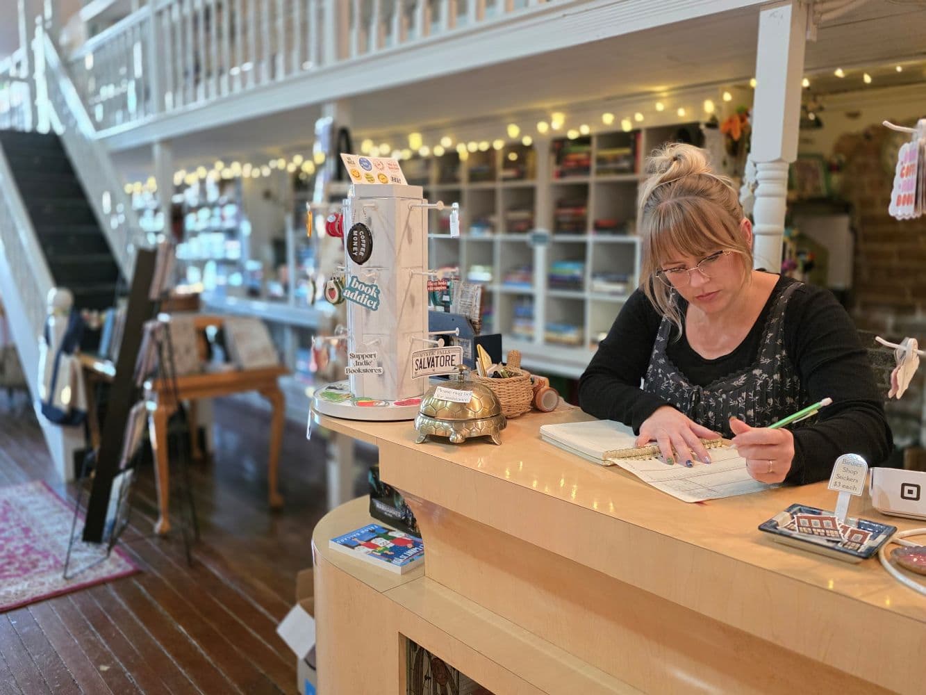 A woman writes notes at a counter in a cozy shop filled with crafts and decor.