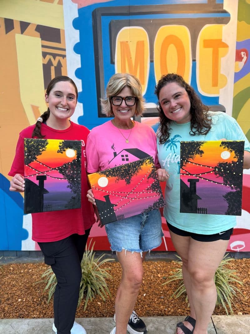 Three women hold painted canvases against a colorful mural backdrop.