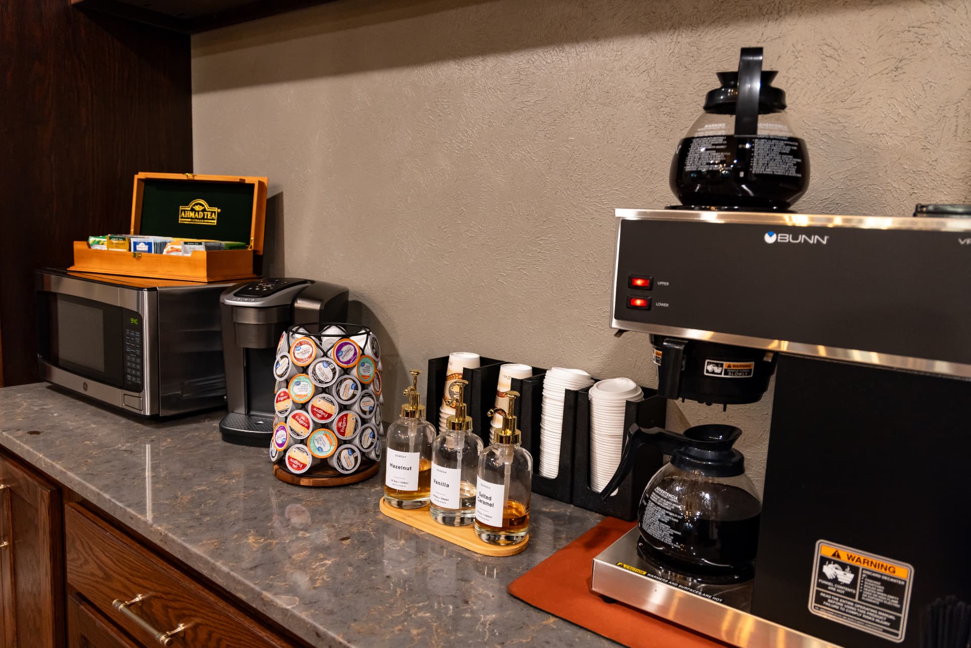 A kitchen countertop featuring a microwave, coffee maker, assortment of coffee pods, and flavored syrups.