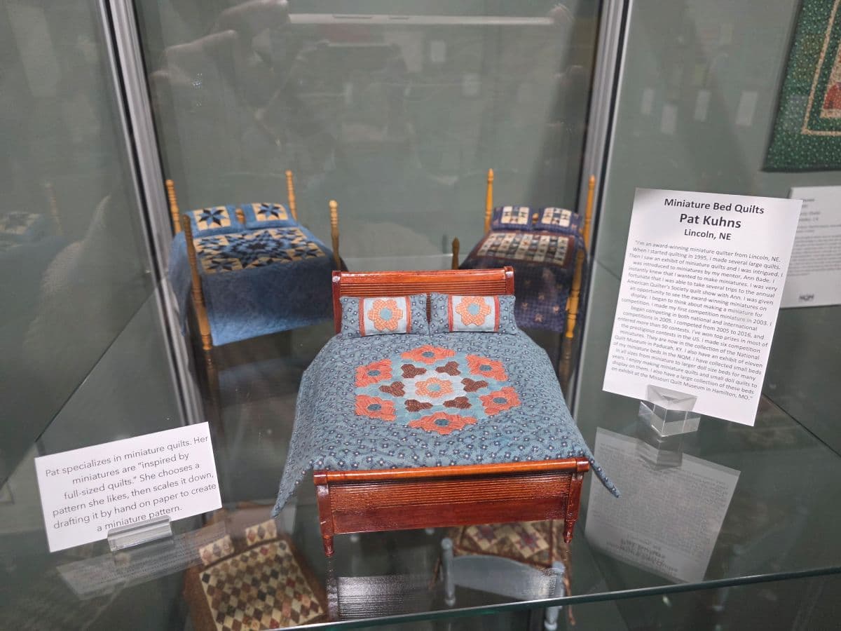 A display case features three miniature bed quilts on tiny beds, with a description of the artist Pat Kuhns.