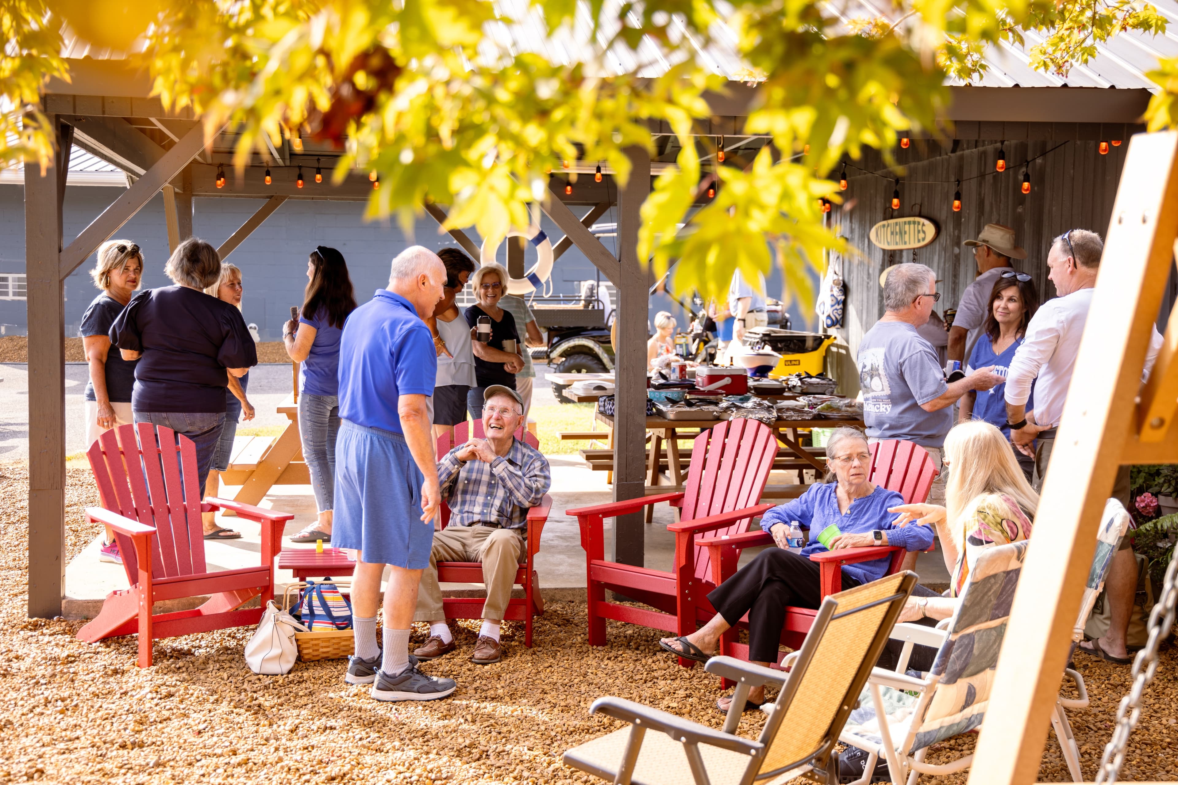 A group of people socialize in a cozy outdoor setting with red Adirondack chairs and a festive atmosphere.