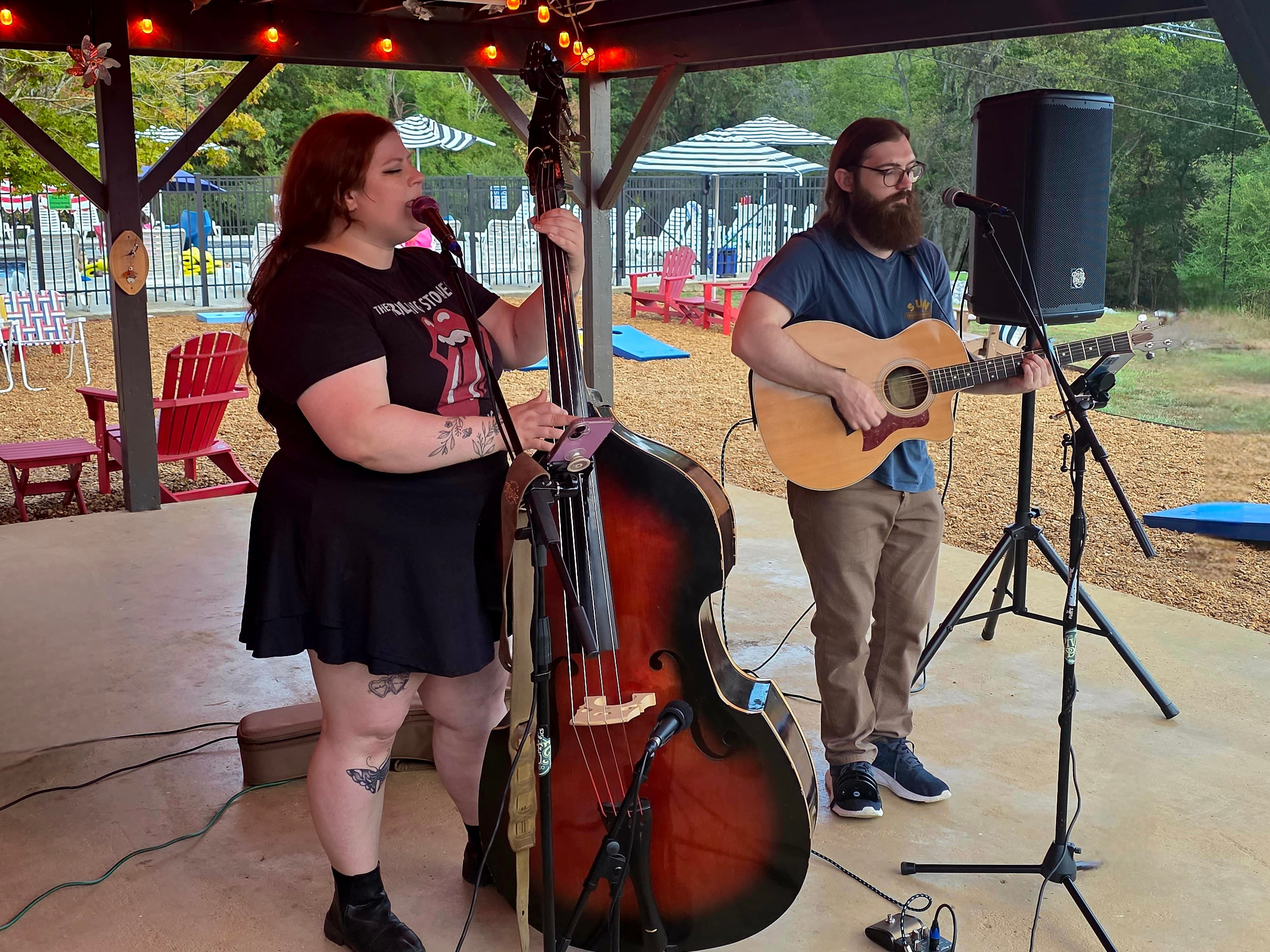 A woman playing a double bass and a man with an acoustic guitar perform music outdoors under a pavilion.