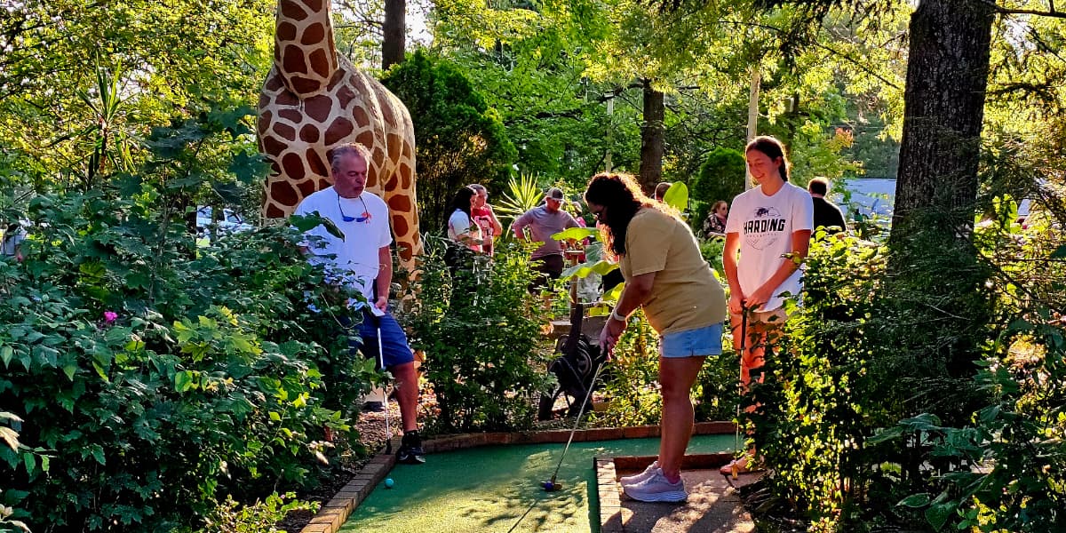 Three people play mini-golf surrounded by greenery and a large giraffe statue.