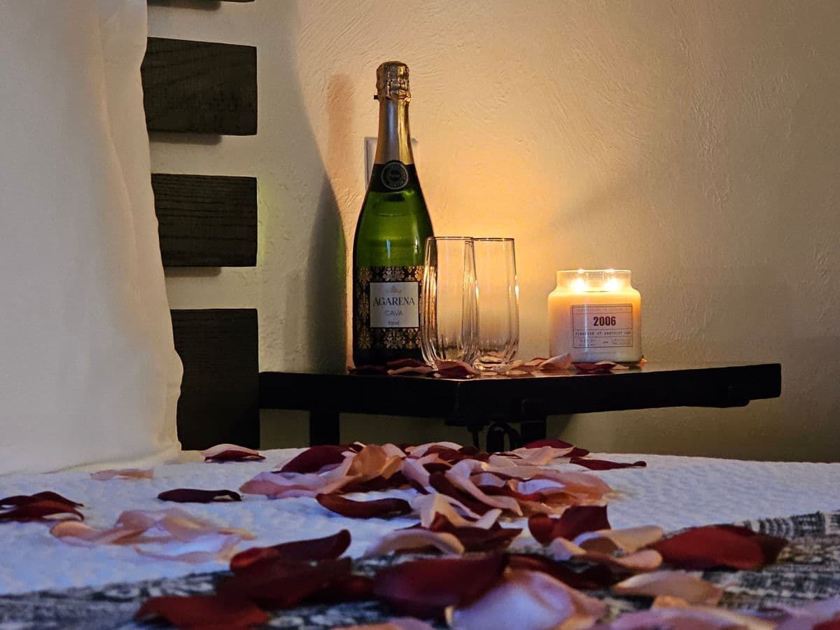 A romantic setup featuring a bottle of champagne, two glasses, a lit candle, and scattered rose petals on a bed.