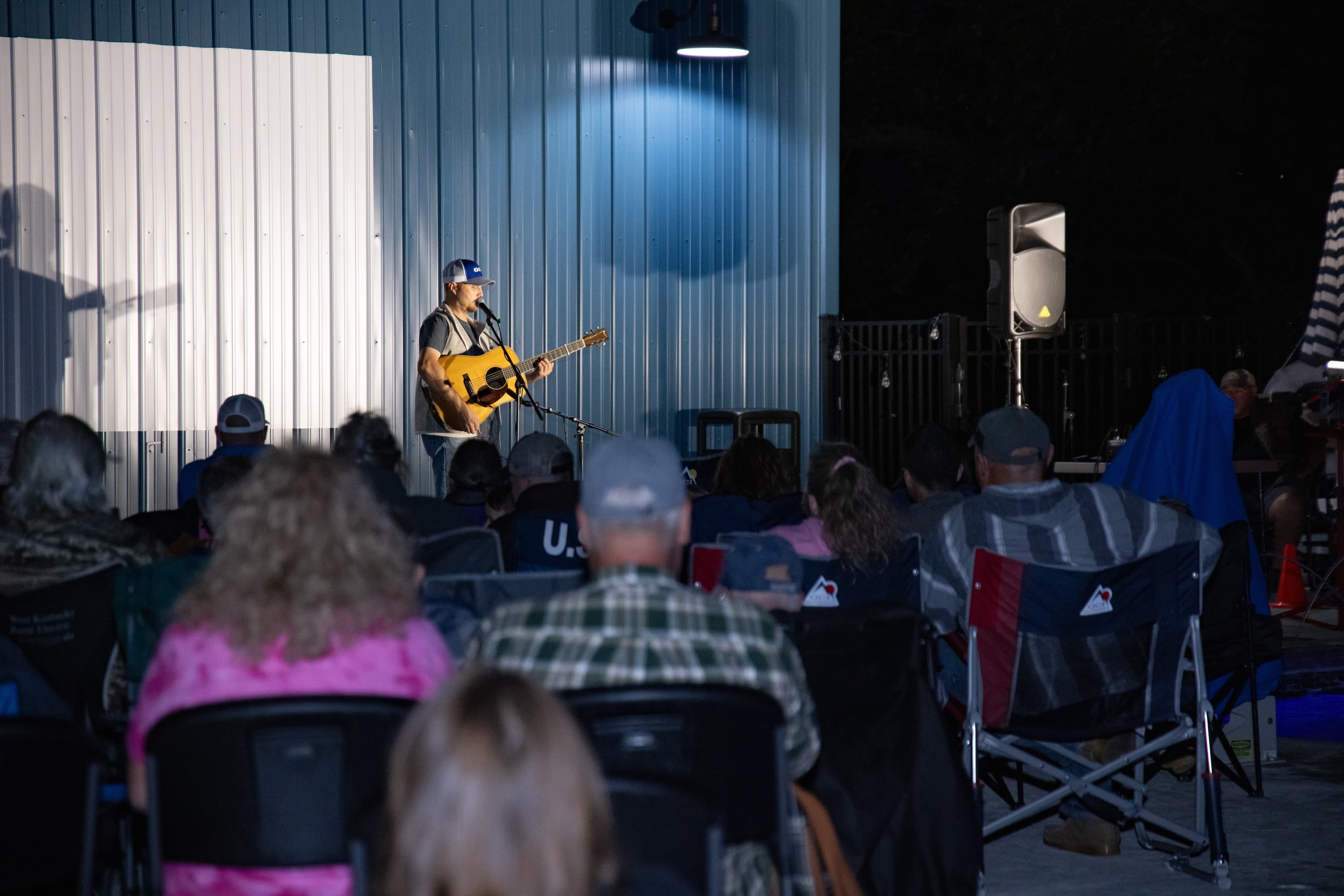 A singer-songwriter performs with a guitar in front of a seated audience at an outdoor venue.