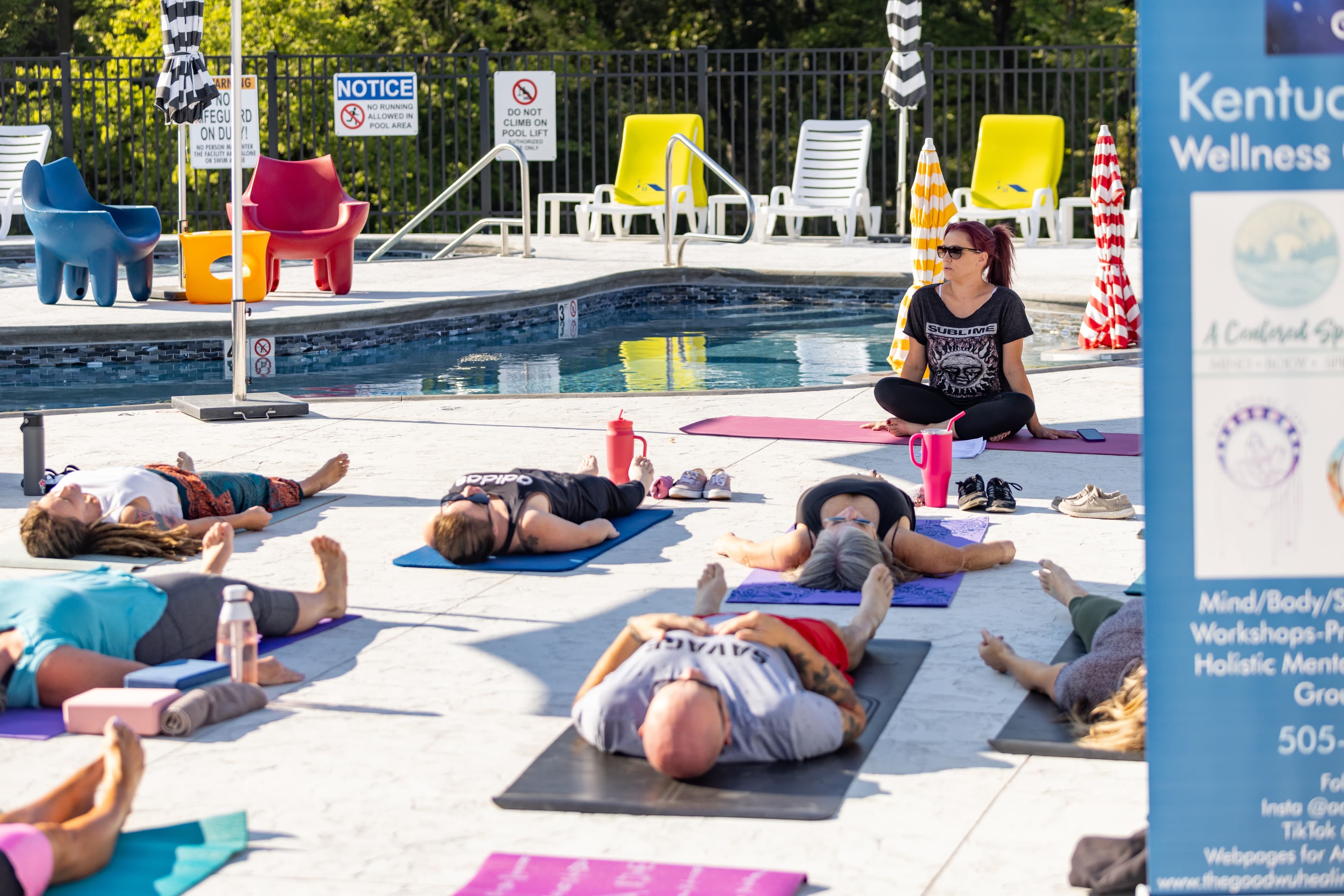 A group of people practice yoga on mats by a pool, while an instructor sits nearby in a sunny outdoor setting.