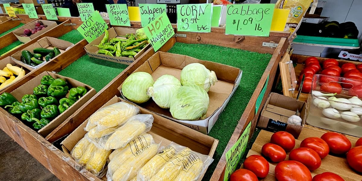 A variety of fresh vegetables, including peppers, corn, cabbage, and tomatoes, displayed in wooden crates with price signs.