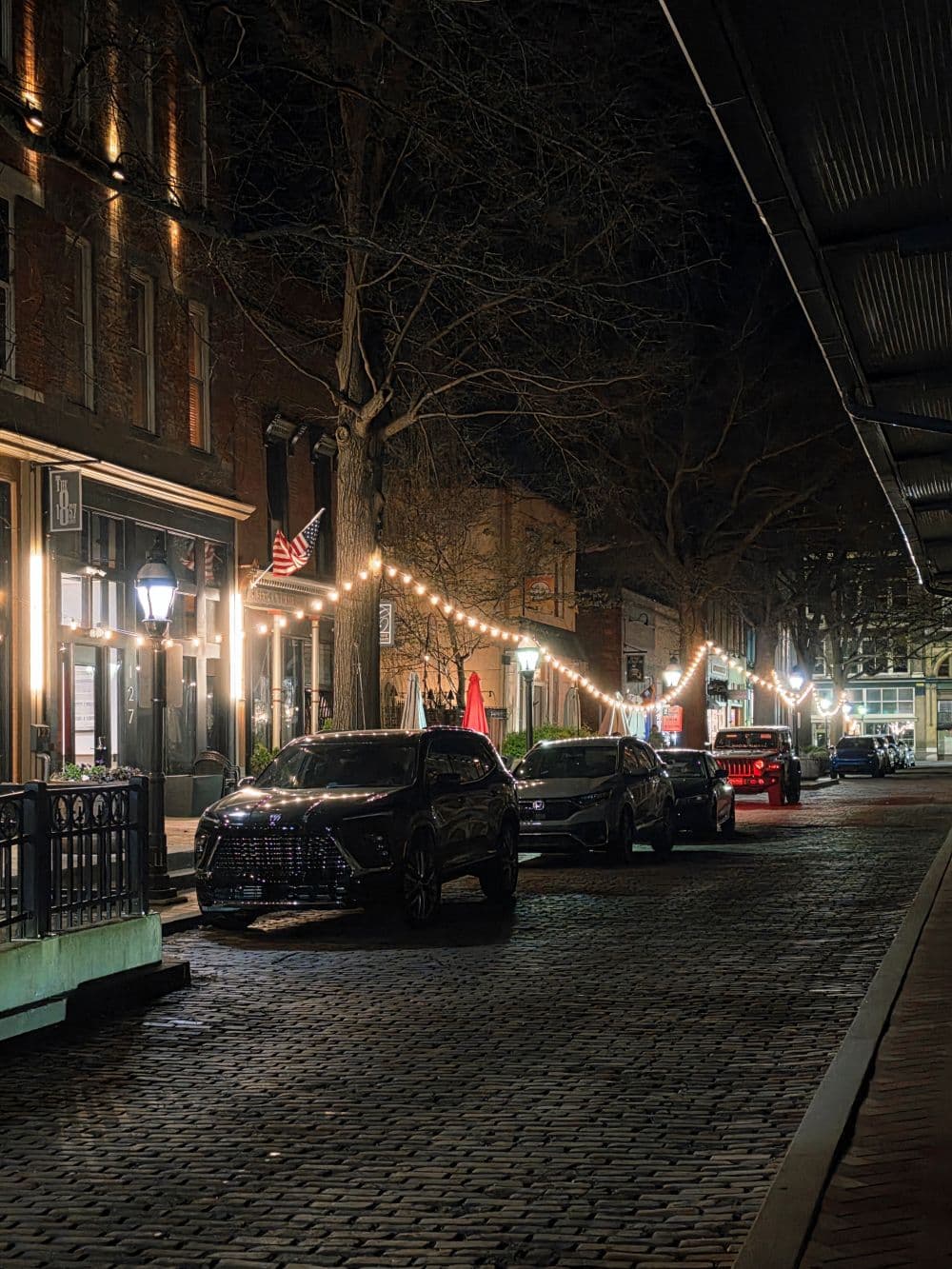 A dimly lit cobblestone street lined with parked cars and adorned with string lights.