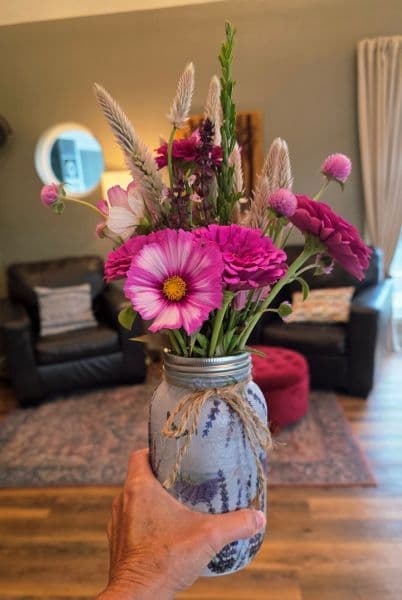 A hand holds a colorful bouquet in a decorated mason jar, set against a cozy living room backdrop.