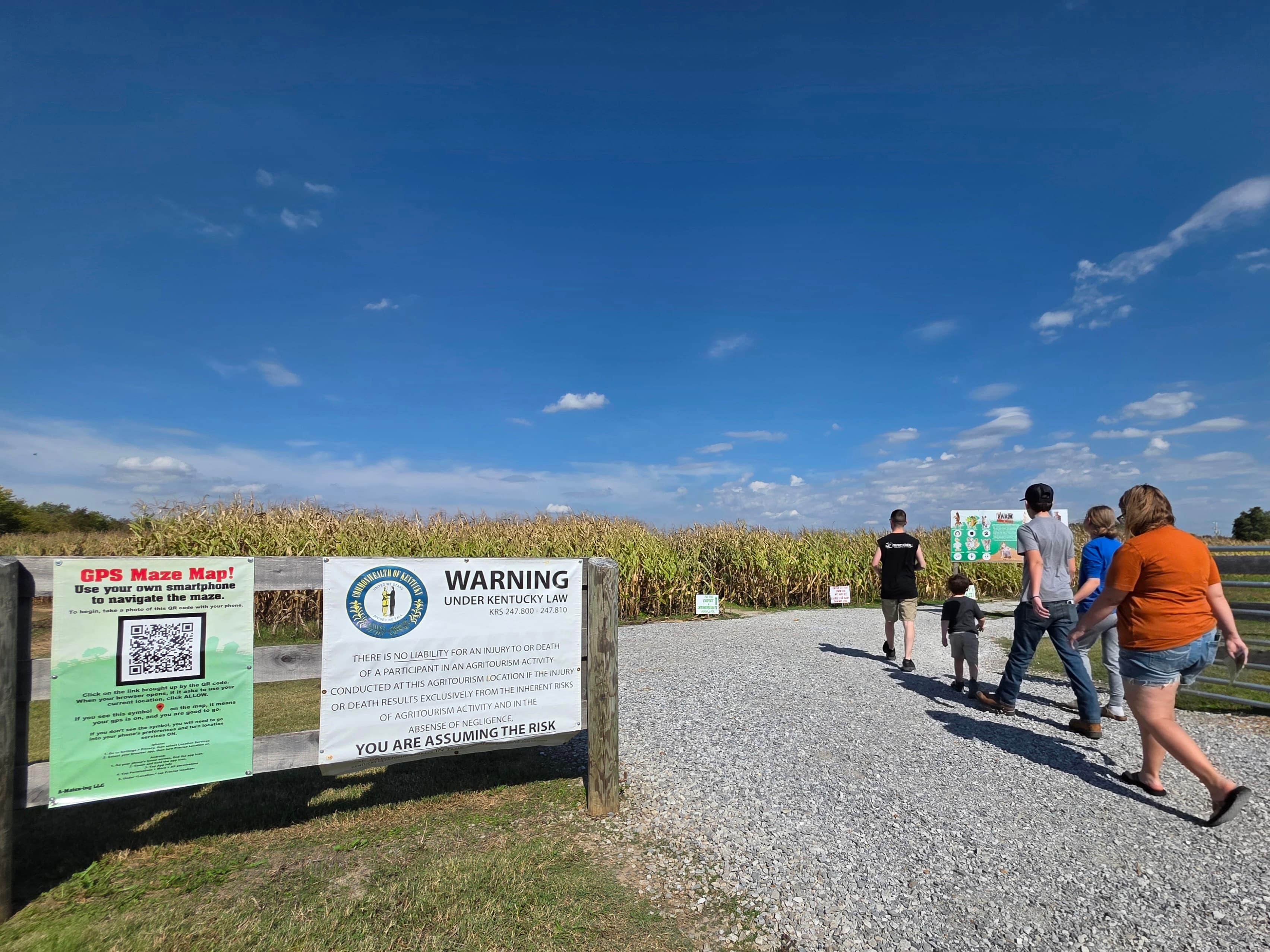 A family walks towards a corn maze entrance with warning signs under a blue sky.