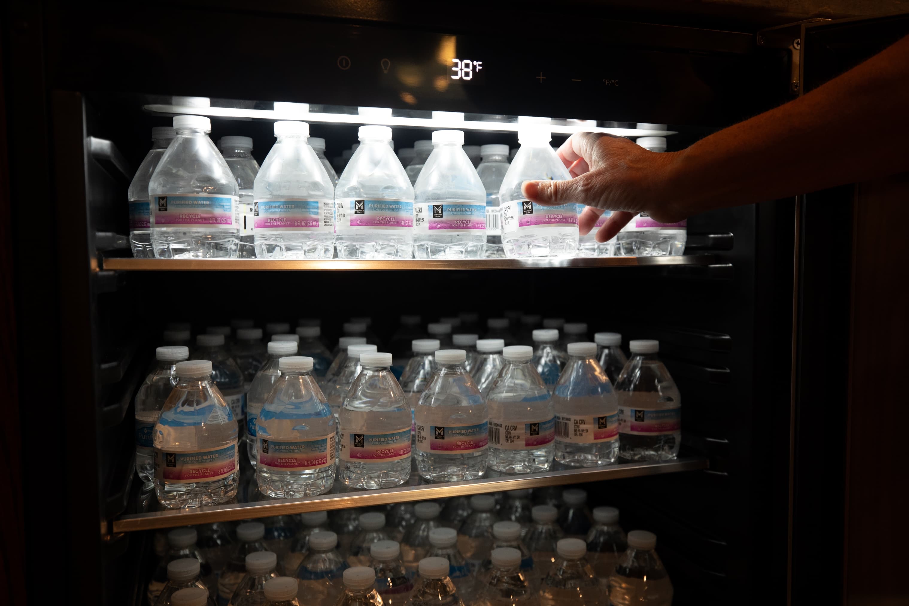 A hand reaches for a bottle of water in a refrigerator filled with water bottles.