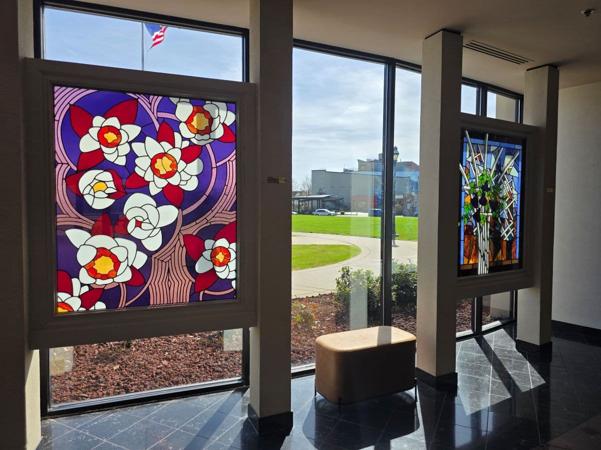 Two stained glass windows with floral designs flank a modern interior space, featuring a view of an outdoor green area through large glass panes.