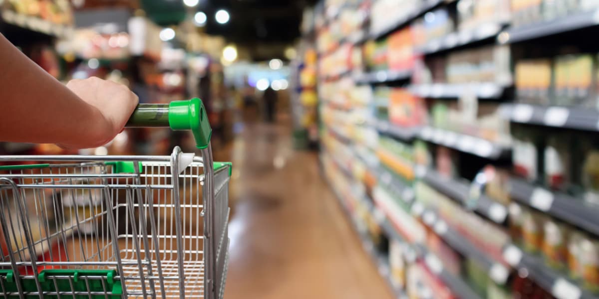 A hand grips the handle of a shopping cart in a grocery aisle filled with shelves of products.