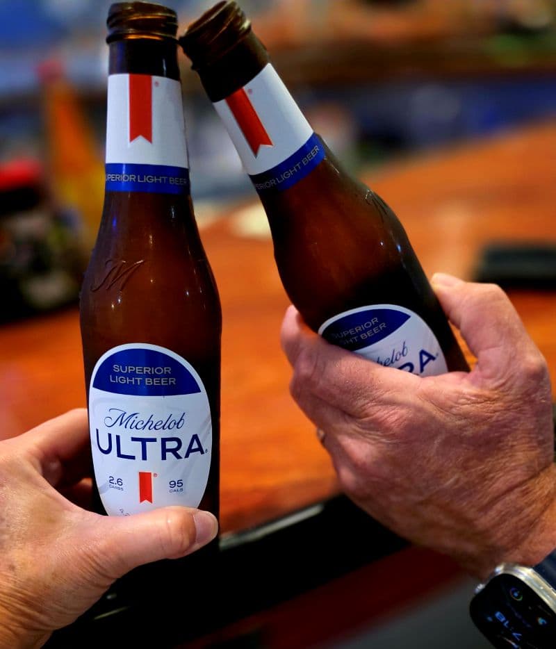 two people off camera clicking together two bottles of Michelob Ultra beer